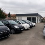 Rangée de voitures citadines compactes garées sur un parking devant un concessionnaire automobile sous un ciel gris.